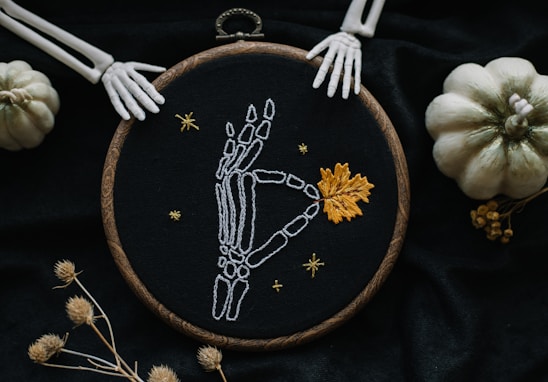 An embroidery hoop displaying a white skeleton hand reaching toward an orange leaf, surrounded by small stars on a black fabric background. The hoop is adorned with an intricate wooden pattern. Two small white pumpkins and skeletal hands frame the hoop, set against a dark, textured cloth. Dried flowers are artistically arranged nearby, enhancing the autumnal theme.