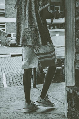 A person wearing a casual outfit including a loose shirt and patterned shorts is captured from the back, standing on a street. The scene includes a parked car and a cobblestone pavement, with a building in the background. The photo has a vintage or muted color tone.