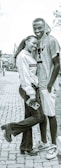 A young couple stands closely together on a paved path, with the girl leaning against the boy and holding a phone. She wears a white jacket and dark jeans, while he sports a gray hoodie and shorts. Both are smiling warmly.