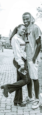 A young couple stands closely together on a paved path, with the girl leaning against the boy and holding a phone. She wears a white jacket and dark jeans, while he sports a gray hoodie and shorts. Both are smiling warmly.