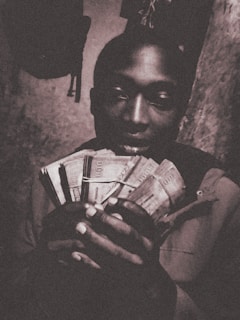 A person is holding a large amount of paper currency in a dimly lit environment. The image has a sepia tone, giving it an aged, vintage appearance. The focus is on the bundle of money prominently displayed in the person's hands.