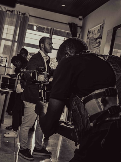 A black and white photograph captures a scene of a cameraman in the foreground holding a professional camera, focusing on a subject. The background includes a few other people, including a man in a suit and others who appear to be conversing or observing.