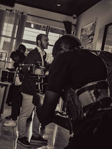 A black and white photograph captures a scene of a cameraman in the foreground holding a professional camera, focusing on a subject. The background includes a few other people, including a man in a suit and others who appear to be conversing or observing.