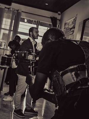 A black and white photograph captures a scene of a cameraman in the foreground holding a professional camera, focusing on a subject. The background includes a few other people, including a man in a suit and others who appear to be conversing or observing.