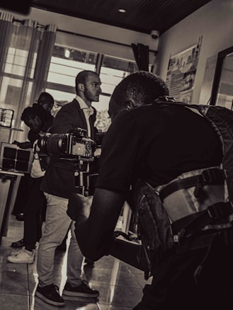 A black and white photograph captures a scene of a cameraman in the foreground holding a professional camera, focusing on a subject. The background includes a few other people, including a man in a suit and others who appear to be conversing or observing.