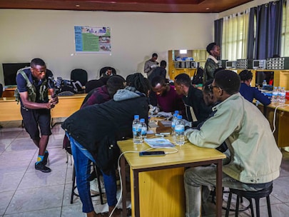 Several people are gathered around a table, appearing to be engaged in a collaborative task or discussion. One person is using a camera on the left side of the image, possibly recording the activity. The room has wooden furniture, a tiled floor, and there are electronic devices and bottles of water on the tables. The atmosphere seems focused and productive.