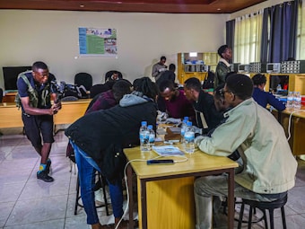 Several people are gathered around a table, appearing to be engaged in a collaborative task or discussion. One person is using a camera on the left side of the image, possibly recording the activity. The room has wooden furniture, a tiled floor, and there are electronic devices and bottles of water on the tables. The atmosphere seems focused and productive.