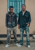 Two men are standing side by side in casual clothing in front of a building. The man on the left wears a sheer black shirt and camouflage pants, with white sneakers. The man on the right wears a dark shirt and distressed jeans, also with white sneakers. Both have a relaxed posture, with one having a hand in his pocket. A window with blue bars is visible behind them.