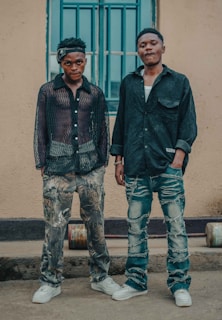 Two men are standing side by side in casual clothing in front of a building. The man on the left wears a sheer black shirt and camouflage pants, with white sneakers. The man on the right wears a dark shirt and distressed jeans, also with white sneakers. Both have a relaxed posture, with one having a hand in his pocket. A window with blue bars is visible behind them.