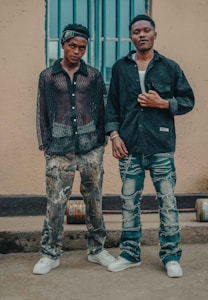 Two young men standing beside each other against a beige wall and a blue-barred window. They are wearing casual, stylish clothing, with one in a sheer black shirt and camouflage pants, and the other in a dark shirt and ripped jeans.