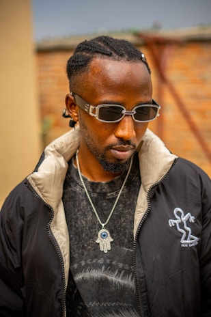 A person with braided hair wearing dark sunglasses, a black jacket with a unique logo, and a T-shirt with a distinct pattern. A necklace featuring a Hamsa symbol with an eye adorns the neck. The setting appears to be outdoors with a brick wall and indistinct background elements.