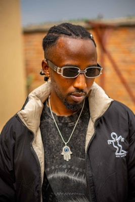 A person with braided hair wearing dark sunglasses, a black jacket with a unique logo, and a T-shirt with a distinct pattern. A necklace featuring a Hamsa symbol with an eye adorns the neck. The setting appears to be outdoors with a brick wall and indistinct background elements.