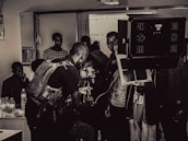 A group of people engaged in a film or photography setting with equipment such as cameras, lights, and a tripod. Some individuals appear to be focused on the technical aspects, possibly setting up or adjusting the gear, while others observe.