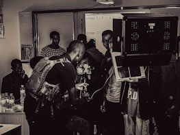 A group of people engaged in a film or photography setting with equipment such as cameras, lights, and a tripod. Some individuals appear to be focused on the technical aspects, possibly setting up or adjusting the gear, while others observe.