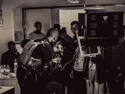 A group of people engaged in a film or photography setting with equipment such as cameras, lights, and a tripod. Some individuals appear to be focused on the technical aspects, possibly setting up or adjusting the gear, while others observe.