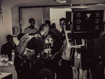 A group of people engaged in a film or photography setting with equipment such as cameras, lights, and a tripod. Some individuals appear to be focused on the technical aspects, possibly setting up or adjusting the gear, while others observe.