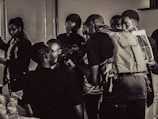 A group of people gathers around an individual holding a camera in a room. The scene is in black and white, giving it a vintage feel. There are bottles on the table and one person appears to be looking at a photo or document. The interaction suggests a workshop or educational setting.