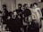 A group of people gathers around an individual holding a camera in a room. The scene is in black and white, giving it a vintage feel. There are bottles on the table and one person appears to be looking at a photo or document. The interaction suggests a workshop or educational setting.