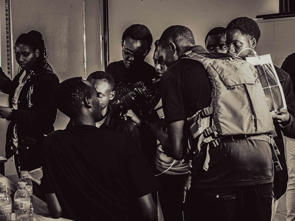 A group of people gathers around an individual holding a camera in a room. The scene is in black and white, giving it a vintage feel. There are bottles on the table and one person appears to be looking at a photo or document. The interaction suggests a workshop or educational setting.