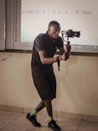 A person is holding a gimbal and camera setup, possibly recording or taking photos, in a room that appears to be a classroom or meeting room. They are wearing a black outfit, colorful socks with smiley faces, and black shoes. There is a projection screen in the background displaying some equations.