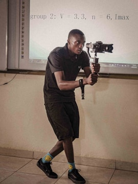 A person is holding a gimbal and camera setup, possibly recording or taking photos, in a room that appears to be a classroom or meeting room. They are wearing a black outfit, colorful socks with smiley faces, and black shoes. There is a projection screen in the background displaying some equations.