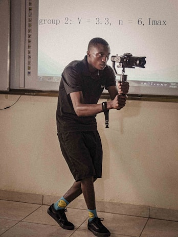 A person is holding a gimbal and camera setup, possibly recording or taking photos, in a room that appears to be a classroom or meeting room. They are wearing a black outfit, colorful socks with smiley faces, and black shoes. There is a projection screen in the background displaying some equations.