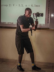 A person in a dark outfit is holding a camera gimbal while standing in a room with a tiled floor. Behind them, a projection on the wall displays equations and textual information. The individual is focused on capturing video or pictures, and the lighting suggests an indoor setting.