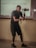 A person in a dark outfit is holding a camera gimbal while standing in a room with a tiled floor. Behind them, a projection on the wall displays equations and textual information. The individual is focused on capturing video or pictures, and the lighting suggests an indoor setting.
