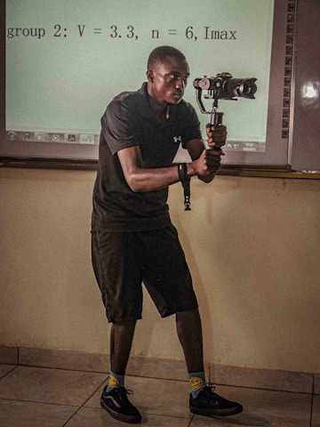 A person in a dark outfit is holding a camera gimbal while standing in a room with a tiled floor. Behind them, a projection on the wall displays equations and textual information. The individual is focused on capturing video or pictures, and the lighting suggests an indoor setting.
