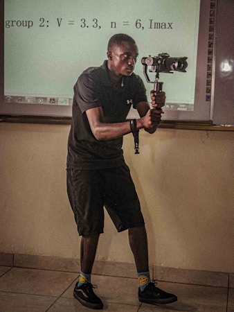 A person in a dark outfit is holding a camera gimbal while standing in a room with a tiled floor. Behind them, a projection on the wall displays equations and textual information. The individual is focused on capturing video or pictures, and the lighting suggests an indoor setting.