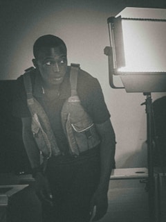 A person wearing a tactical vest is standing beside a bright studio light, casting a dramatic shadow. The image has a monochromatic color scheme, giving it a noir or surveillance aesthetic.