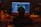 A person seated in a dimly lit room is working at a desk with a large computer screen displaying music production software. Various equipment, including speakers, an audio interface, and a drink bottle labeled 'HO.COOL', are visible. A bottle labeled 'Red Grape' is also on the desk.
