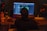 A person seated in a dimly lit room is working at a desk with a large computer screen displaying music production software. Various equipment, including speakers, an audio interface, and a drink bottle labeled 'HO.COOL', are visible. A bottle labeled 'Red Grape' is also on the desk.