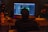 A person seated in a dimly lit room is working at a desk with a large computer screen displaying music production software. Various equipment, including speakers, an audio interface, and a drink bottle labeled 'HO.COOL', are visible. A bottle labeled 'Red Grape' is also on the desk.
