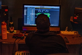 A person seated in a dimly lit room is working at a desk with a large computer screen displaying music production software. Various equipment, including speakers, an audio interface, and a drink bottle labeled 'HO.COOL', are visible. A bottle labeled 'Red Grape' is also on the desk.