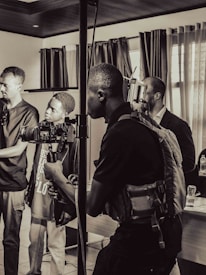 Several people are gathered indoors, focused on filming or recording an event. One person is holding a video camera stabilizer, and others appear to be adjusting equipment or observing the process. The setting includes curtains, tables with various items, and a window letting in natural light.