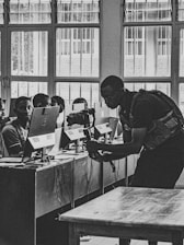 A black and white photograph of a classroom where multiple students are sitting at desks with computers. A person is interacting with the students, possibly teaching or assisting them. Large windows line the wall in the background, allowing natural light to illuminate the room.