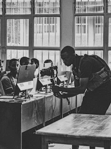 A black and white photograph of a classroom where multiple students are sitting at desks with computers. A person is interacting with the students, possibly teaching or assisting them. Large windows line the wall in the background, allowing natural light to illuminate the room.
