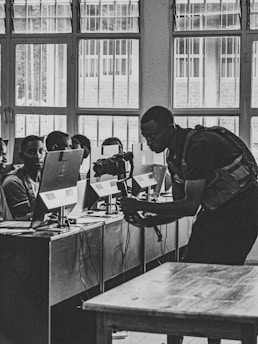 A black and white photograph of a classroom where multiple students are sitting at desks with computers. A person is interacting with the students, possibly teaching or assisting them. Large windows line the wall in the background, allowing natural light to illuminate the room.