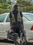 A person stands next to a white car on a paved area, holding professional video equipment including a camera rig and a monitor. The individual is wearing a dark outfit with a utility vest. Greenery is visible in the background, adding a touch of nature to the scene.