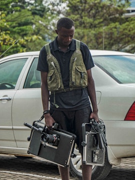 A person stands next to a white car on a paved area, holding professional video equipment including a camera rig and a monitor. The individual is wearing a dark outfit with a utility vest. Greenery is visible in the background, adding a touch of nature to the scene.