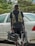A person stands next to a white car on a paved area, holding professional video equipment including a camera rig and a monitor. The individual is wearing a dark outfit with a utility vest. Greenery is visible in the background, adding a touch of nature to the scene.