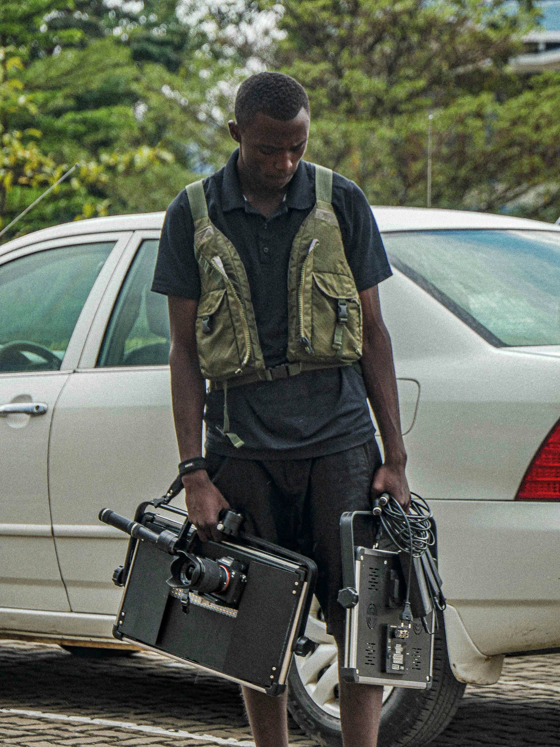 A person stands next to a white car on a paved area, holding professional video equipment including a camera rig and a monitor. The individual is wearing a dark outfit with a utility vest. Greenery is visible in the background, adding a touch of nature to the scene.