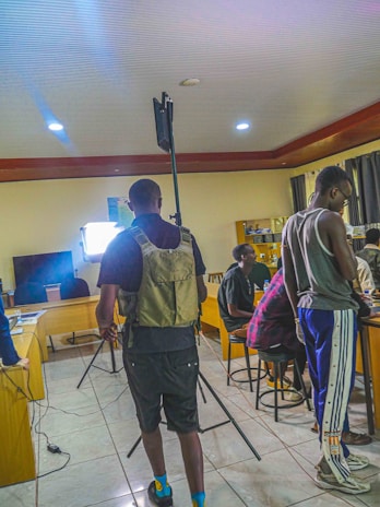A group of people are gathered in a room with camera equipment set up. One person is wearing a tactical vest and seems to be adjusting or preparing equipment. Others are seated or standing casually, and there are desks and chairs in the background along with some electronic devices. The lighting setup suggests some sort of filming or photography session.