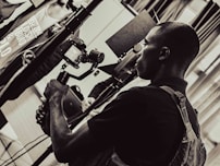 A person is holding a camera mounted on a stabilizer, intently focusing on the viewfinder. The setting appears to be indoors with multiple people and equipment present, giving the scene a professional or documentary atmosphere. The image is in black and white, adding a timeless or classic feel.