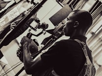 A person is holding a camera mounted on a stabilizer, intently focusing on the viewfinder. The setting appears to be indoors with multiple people and equipment present, giving the scene a professional or documentary atmosphere. The image is in black and white, adding a timeless or classic feel.