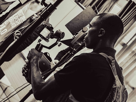 A person is holding a camera mounted on a stabilizer, intently focusing on the viewfinder. The setting appears to be indoors with multiple people and equipment present, giving the scene a professional or documentary atmosphere. The image is in black and white, adding a timeless or classic feel.
