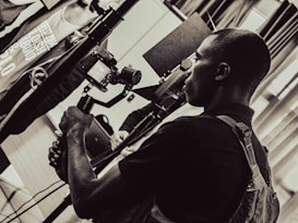 A person is holding a camera mounted on a stabilizer, intently focusing on the viewfinder. The setting appears to be indoors with multiple people and equipment present, giving the scene a professional or documentary atmosphere. The image is in black and white, adding a timeless or classic feel.