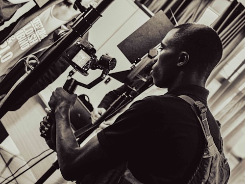 A person is holding a camera mounted on a stabilizer, intently focusing on the viewfinder. The setting appears to be indoors with multiple people and equipment present, giving the scene a professional or documentary atmosphere. The image is in black and white, adding a timeless or classic feel.