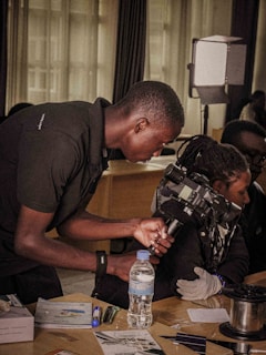 A person is leaning over a table, operating a video camera, with another individual nearby who is wearing gloves. Various objects, including a water bottle, pens, and papers, are scattered on the table. Lighting equipment is visible in the background, suggesting a setup for filming or recording.